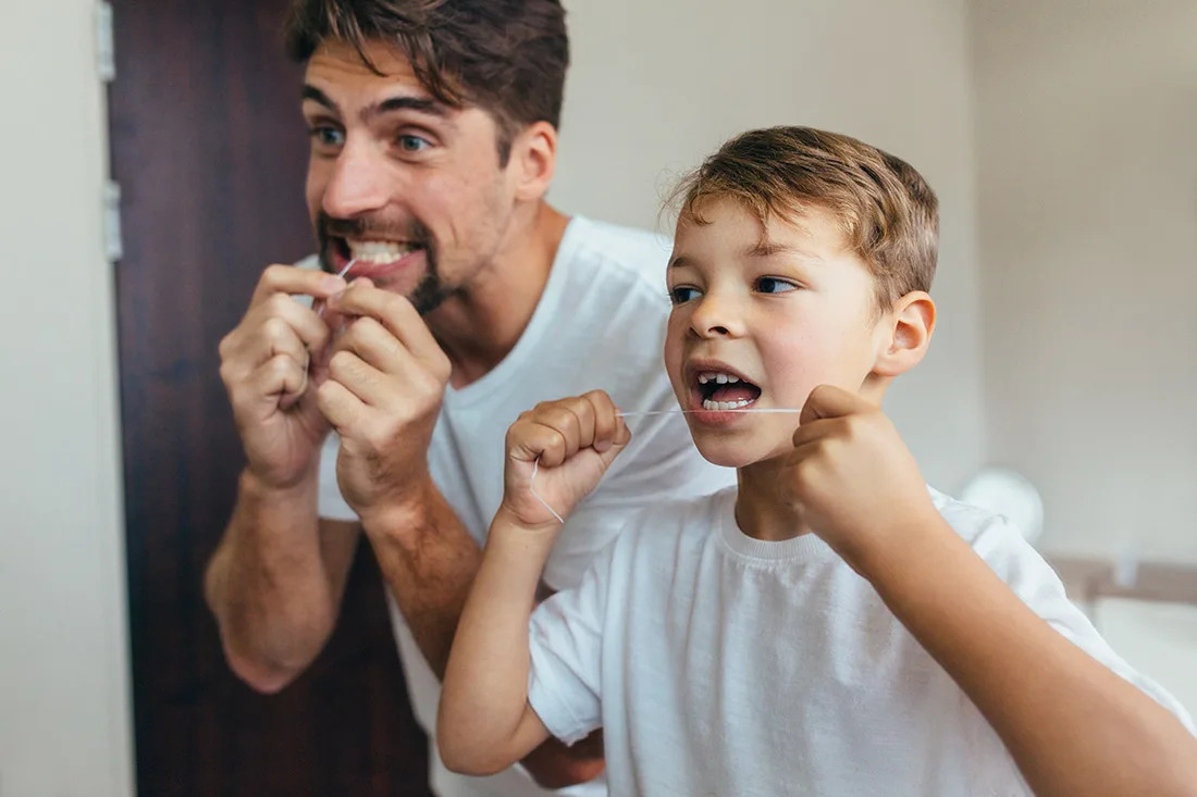 A photo of a parent helping a young child practice proper brushing and flossing habits at home.