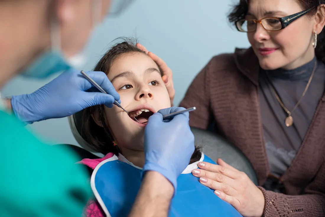 A photo of a pediatric dentist examining a child while a parent offers comfort during an emergency dental visit.
