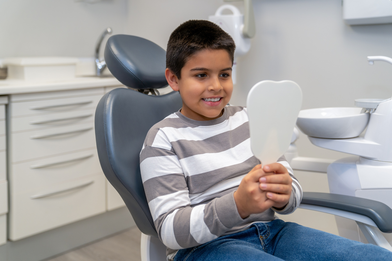 Happy boy sitting in dentist chair, holding a mirror and checking his teeth after dental checkup