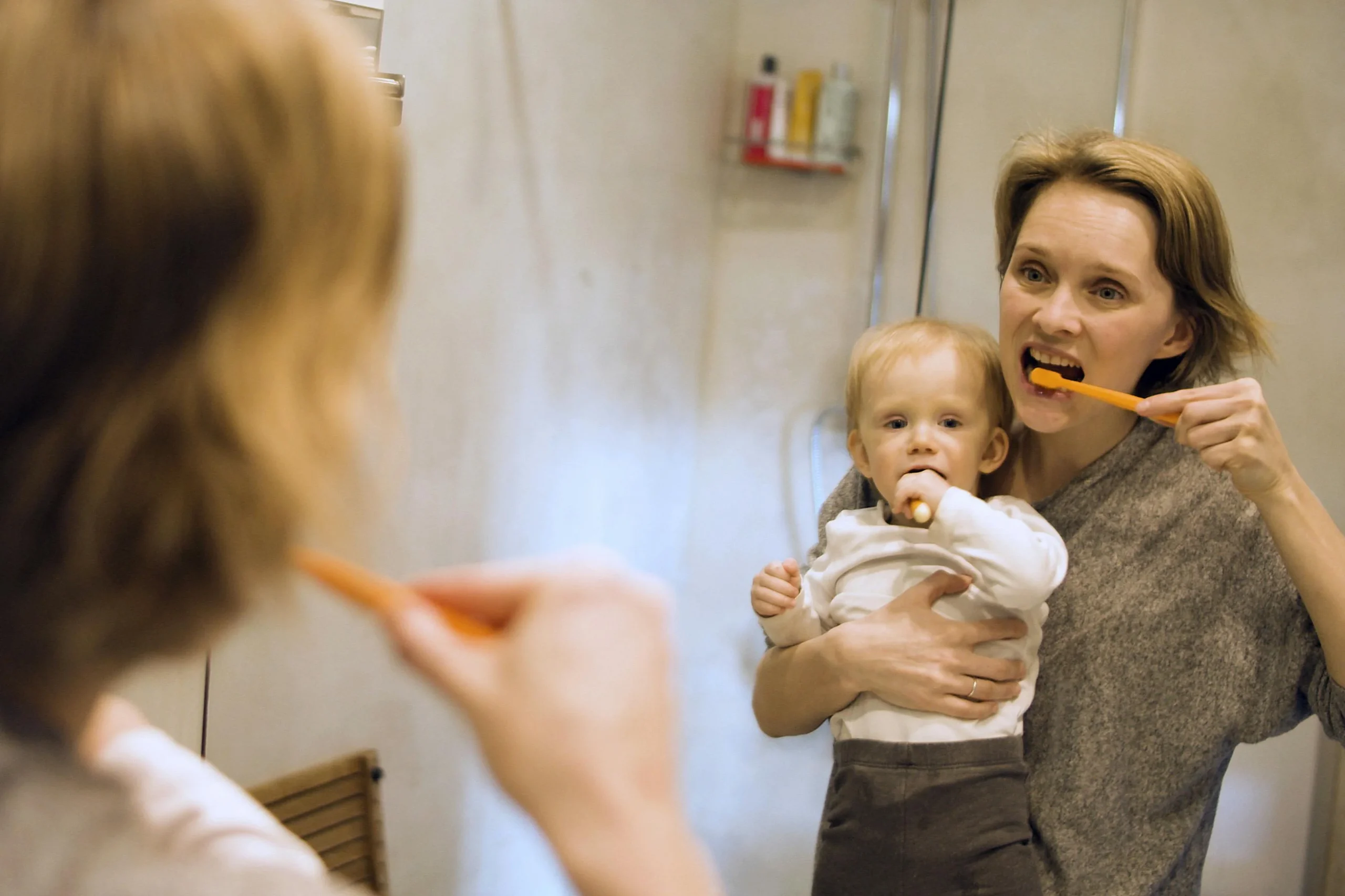 An image of a parent brushing their teeth while holding a baby to model healthy oral hygiene habits at home, showing when do kids start going to the dentist.