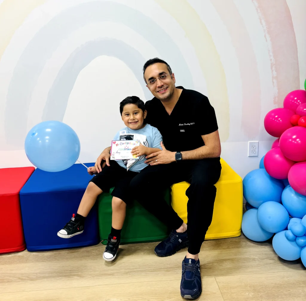 An image of a pediatric dentist and a young child smiling together in a colourful dental clinic, holding a ChildSmiles certificate