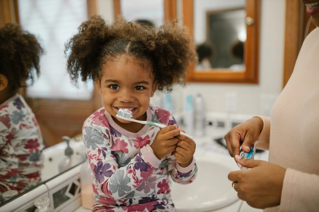 photo of a little girl sitting at a bathroom sink while brushing her teeth with her mom
